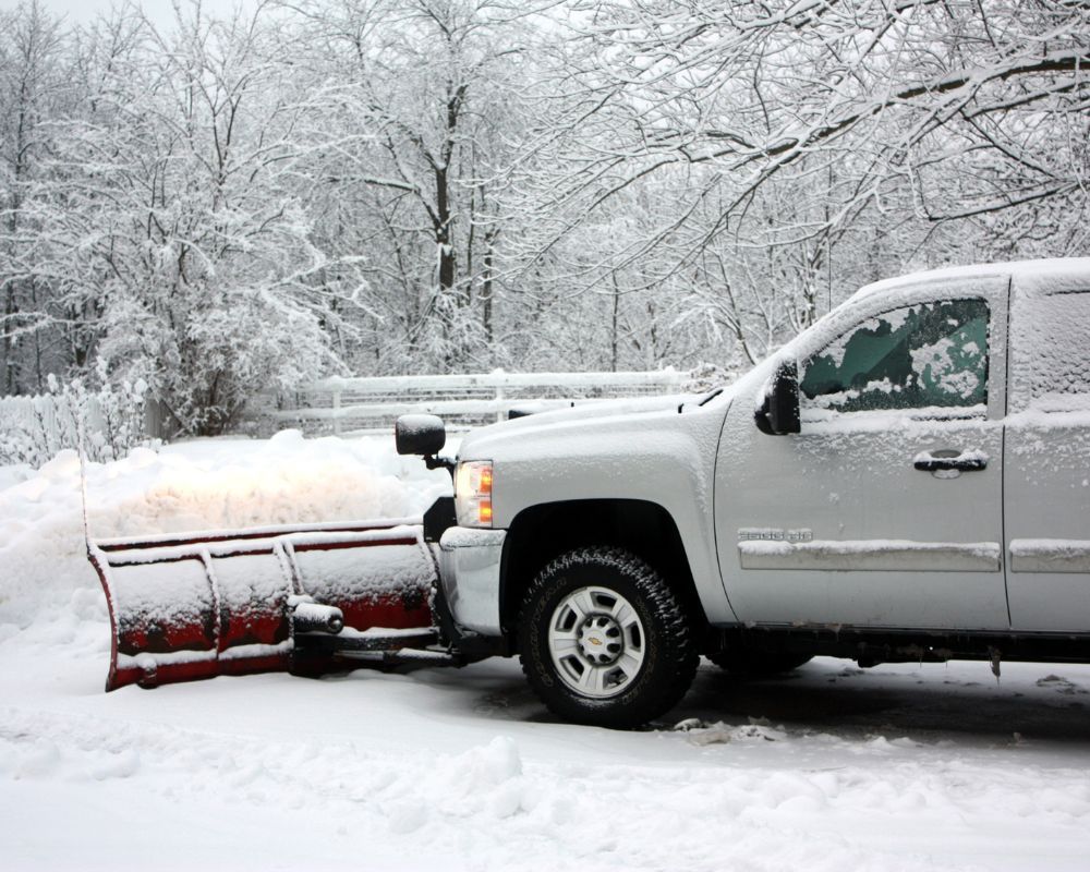 White pickup with red snowplow clearing a snowy road in a winter landscape