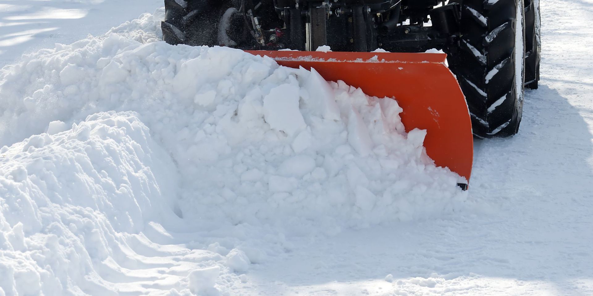 Orange snowplow blade pushing snow beside large tire on a snowy road