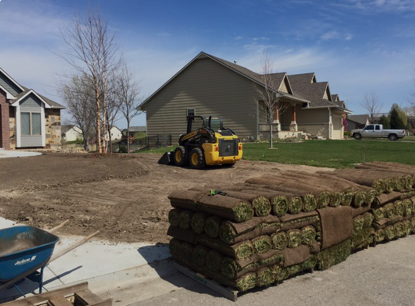 A small bulldozer grading a yard beside stacked sod rolls in a suburban neighborhood.