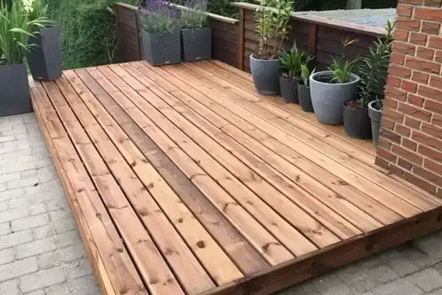 Wooden deck patio with potted plants along a brick wall beside a paved walkway