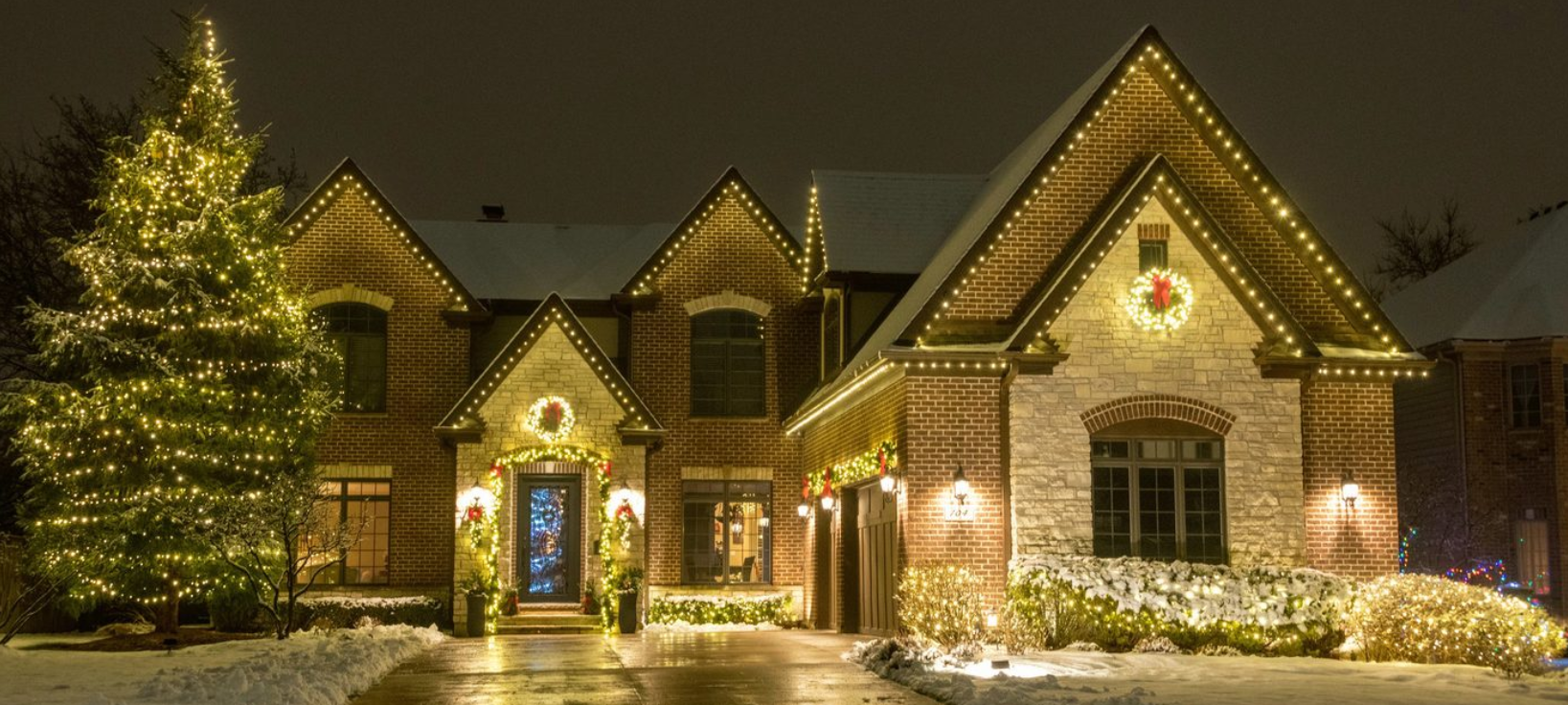 Snow-covered brick house decorated with Christmas lights at night, with a lit tree in front