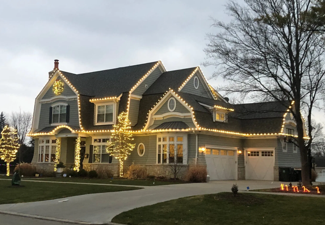 Two-story suburban house at dusk, outlined with warm white Christmas lights and lit windows