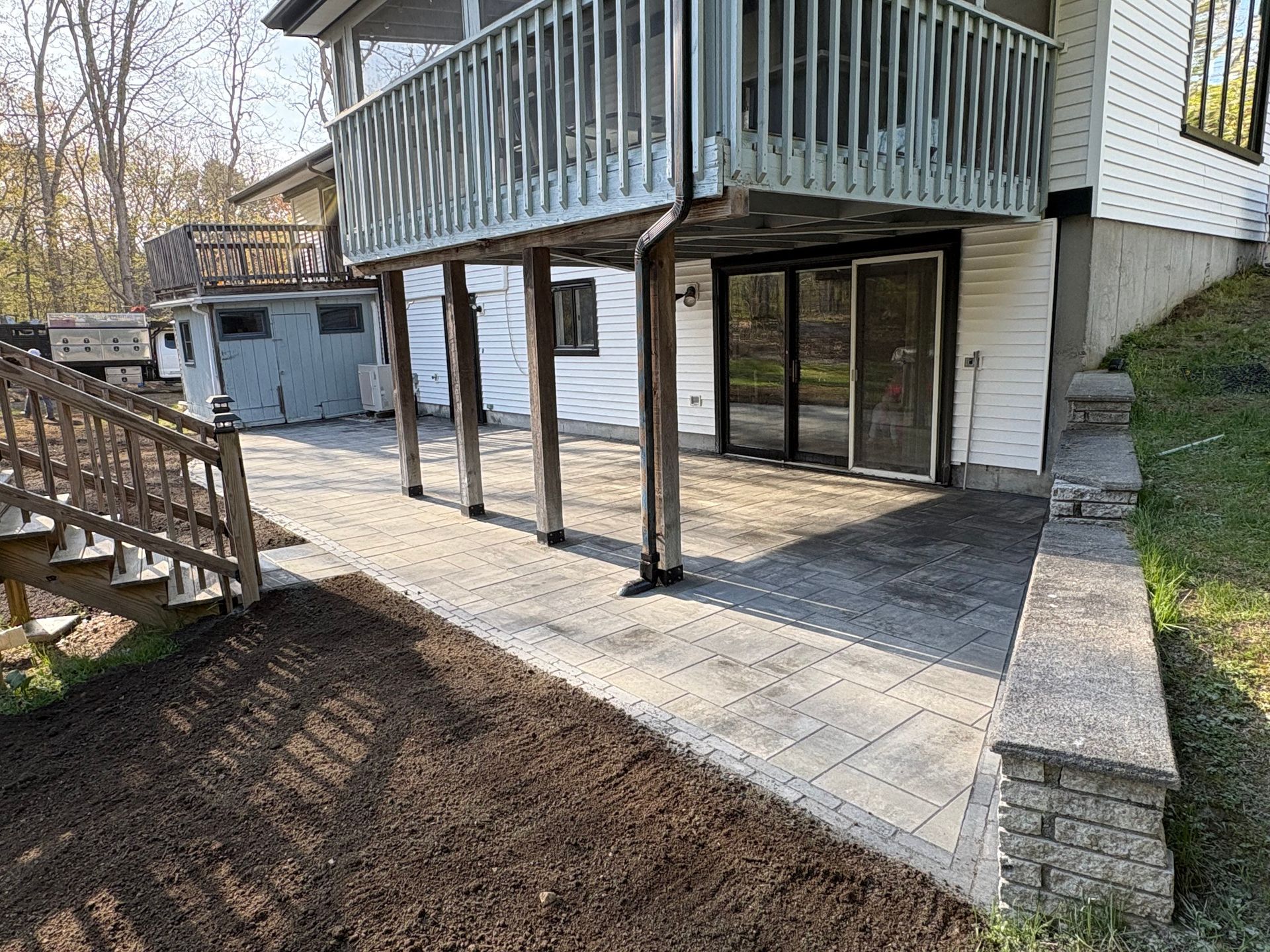 Wooden deck on stilts over a sloped yard, with stairs, patio space, and stone retaining wall.