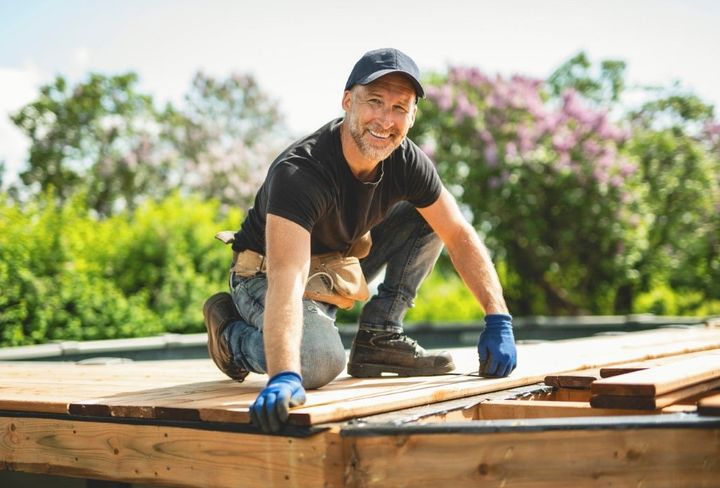 Worker in gloves kneeling on a wooden deck frame outdoors, measuring and fitting planks.