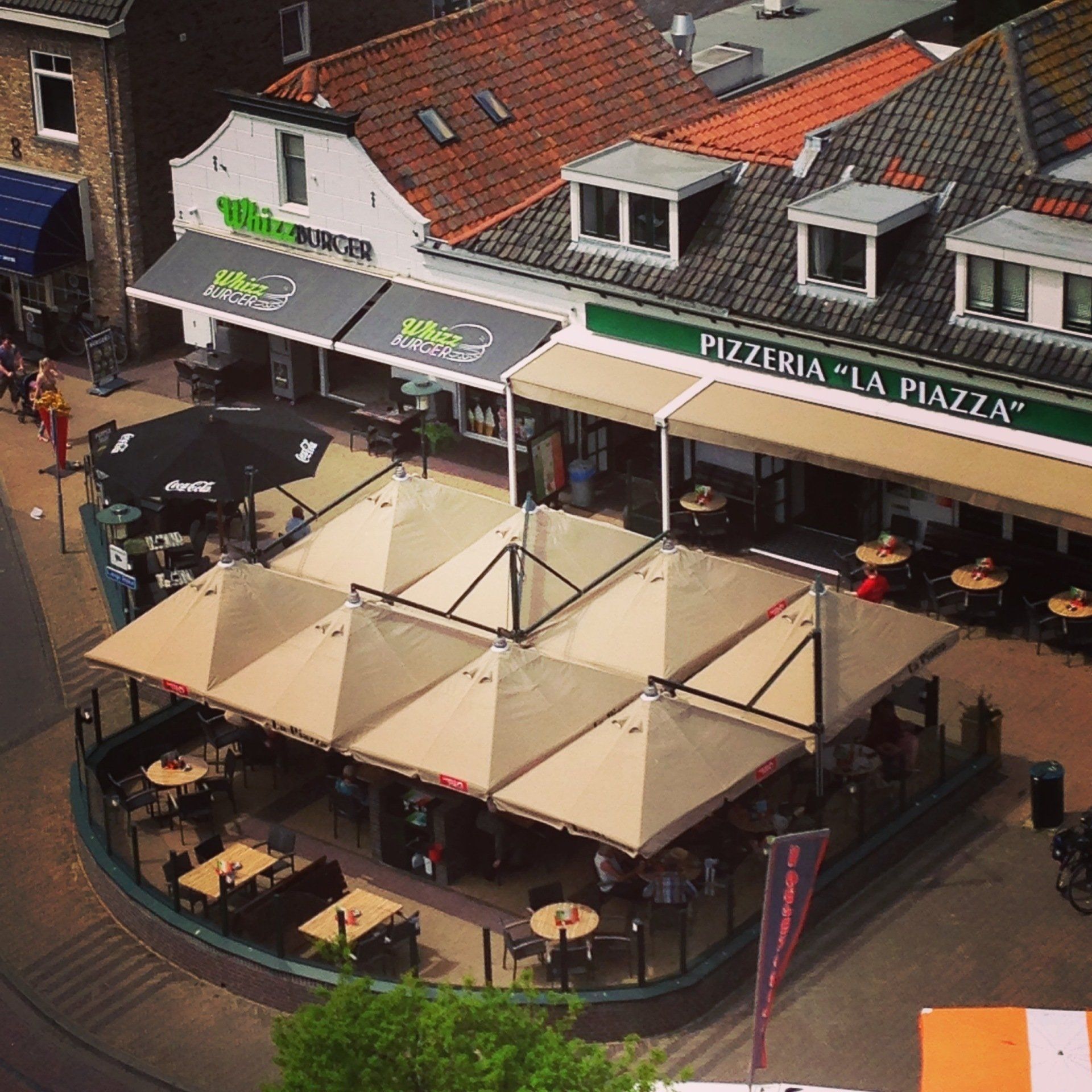 Terras bij Pizzeria La Piazza met crèmekleurige parasols en tafels naast een gebouw met een rood dak.