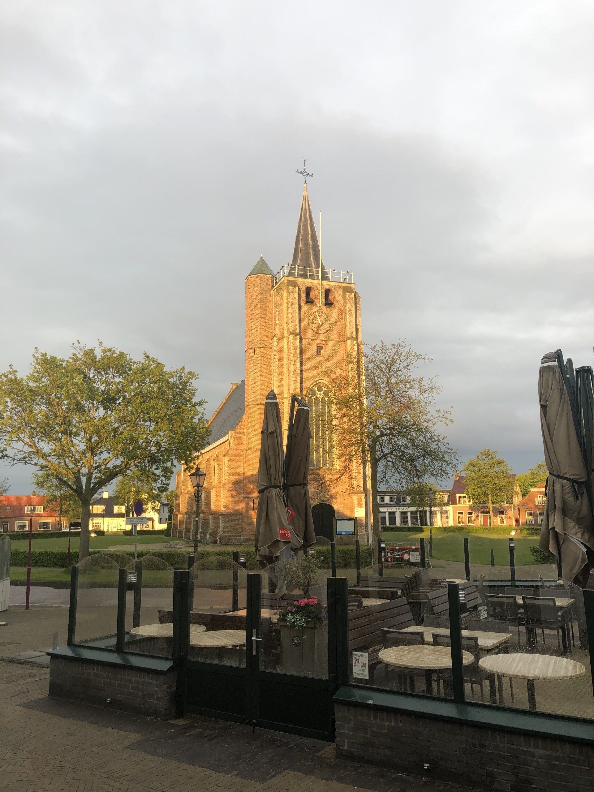 Historische bakstenen kerk met een hoge torenspits, gezien achter een glazen terras van een café op een bewolkte dag.