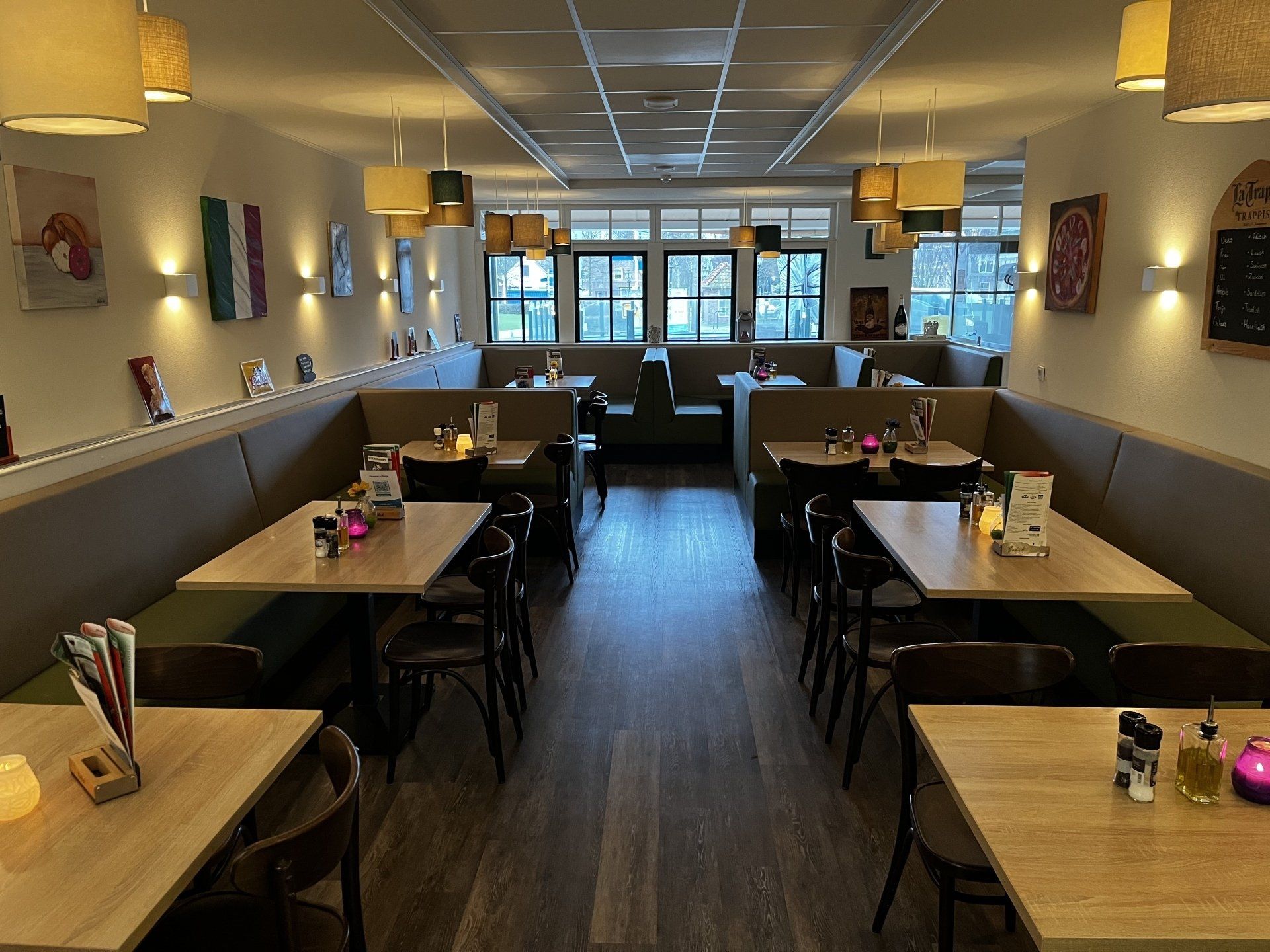 Een lege, moderne restaurantzaal met zitjes in nisjes, hanglampen en tafels bij een raam aan de voorzijde.