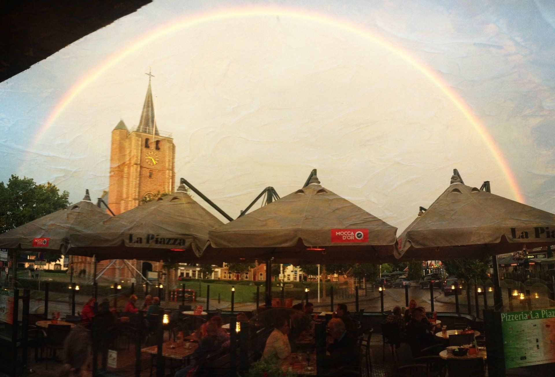 Regenboog boven een terrasje met een kerktoren op de achtergrond bij zonsondergang.