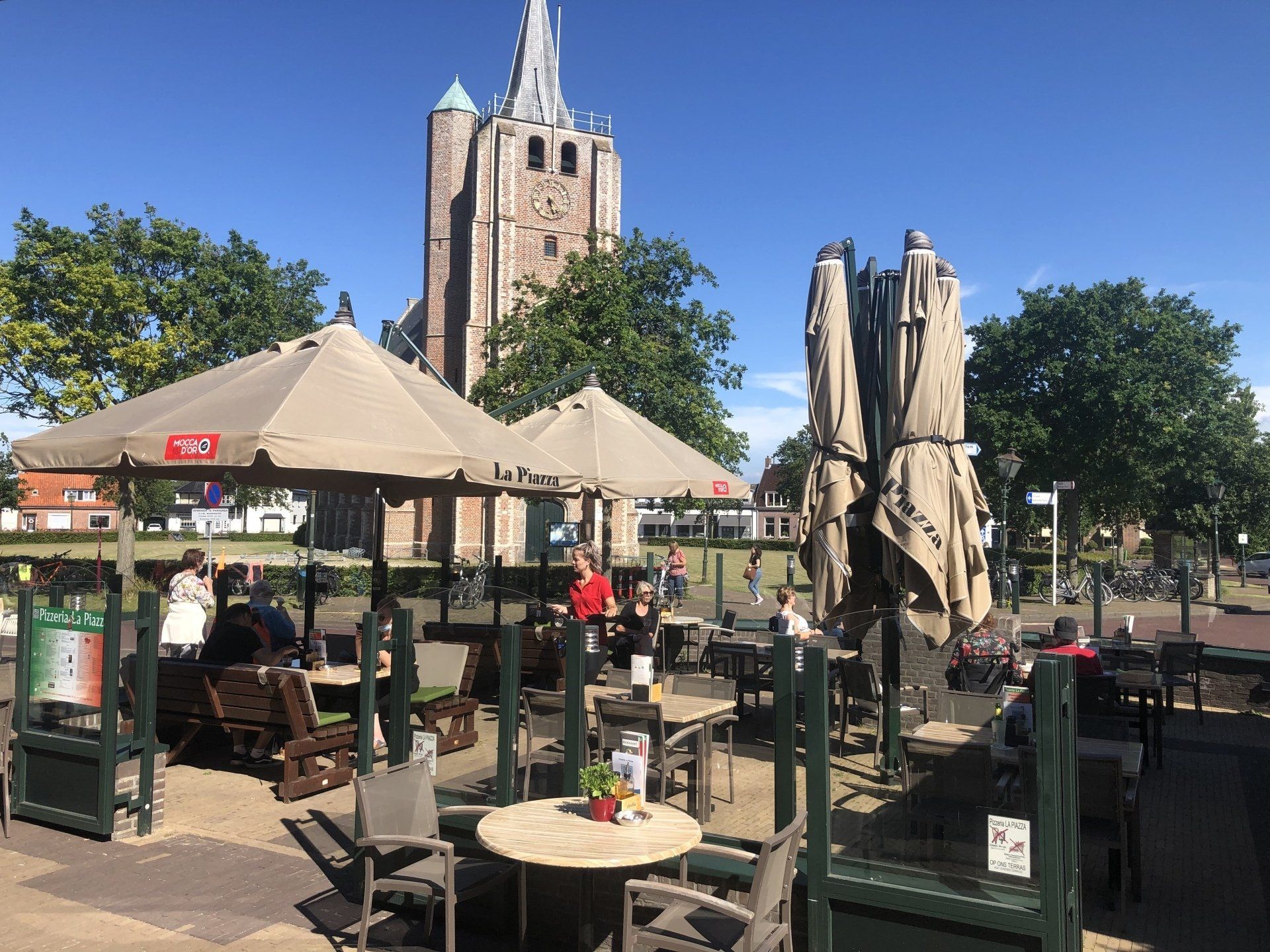 Terras van een café in de buitenlucht met beige parasols en stoelen, met uitzicht op een plein met een kerktoren op de achtergrond.