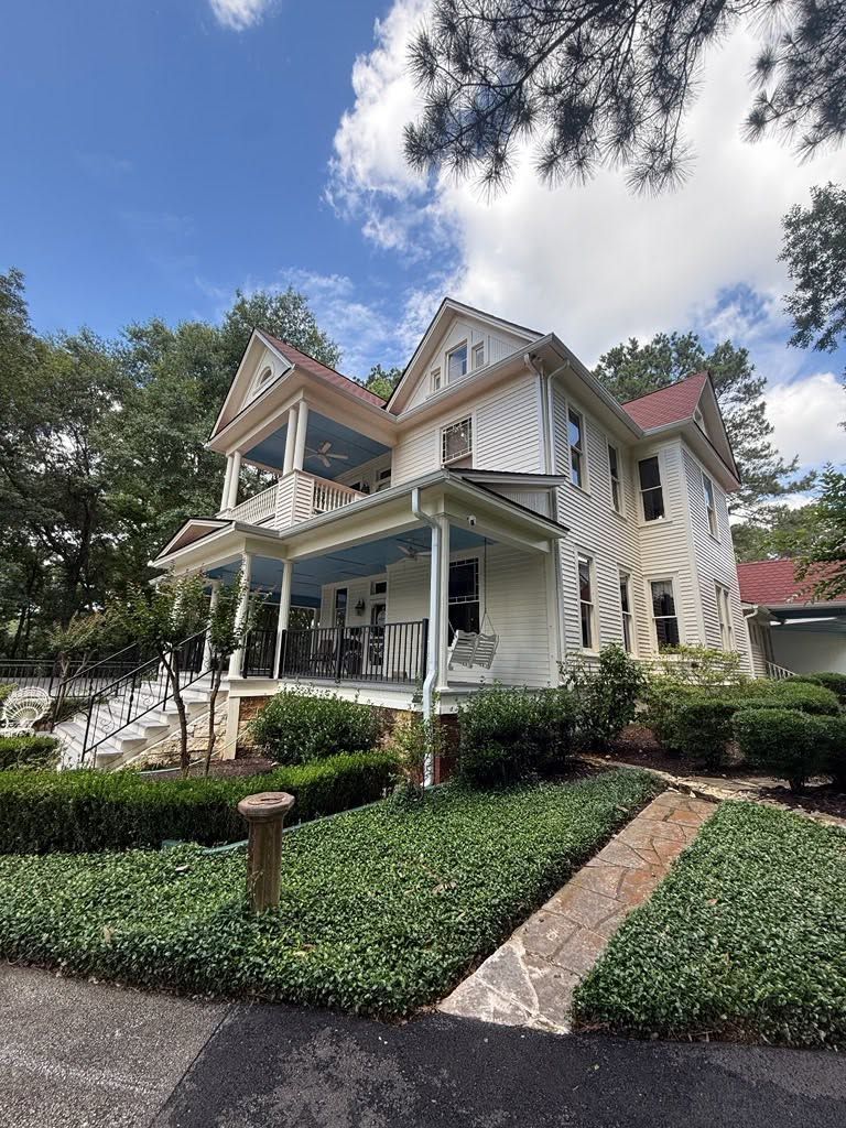 Victorian-style white house with wraparound porch, manicured garden, and tall trees under a partly cloudy sky