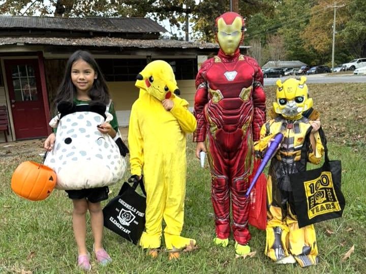 Four children in Halloween costumes posing outdoors with trick-or-treat bags and a pumpkin bucket