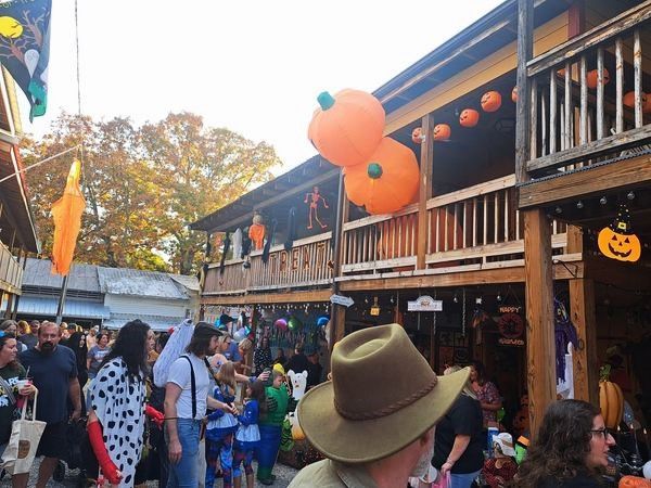 Crowded Halloween street party with costumed people, orange balloons, and pumpkin decorations beside wooden balconies