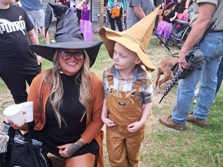 Woman kneels beside child in a straw hat and overalls at a lively outdoor event.