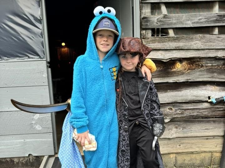 Two children in costumes pose by a rustic wooden wall, one in a blue monster hoodie and one in a dark outfit.