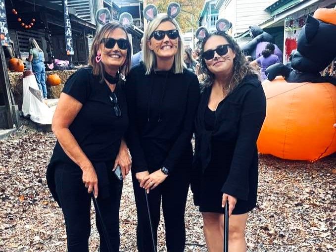 Three people in black outfits pose outdoors at a fall market beside a large orange pumpkin decoration.