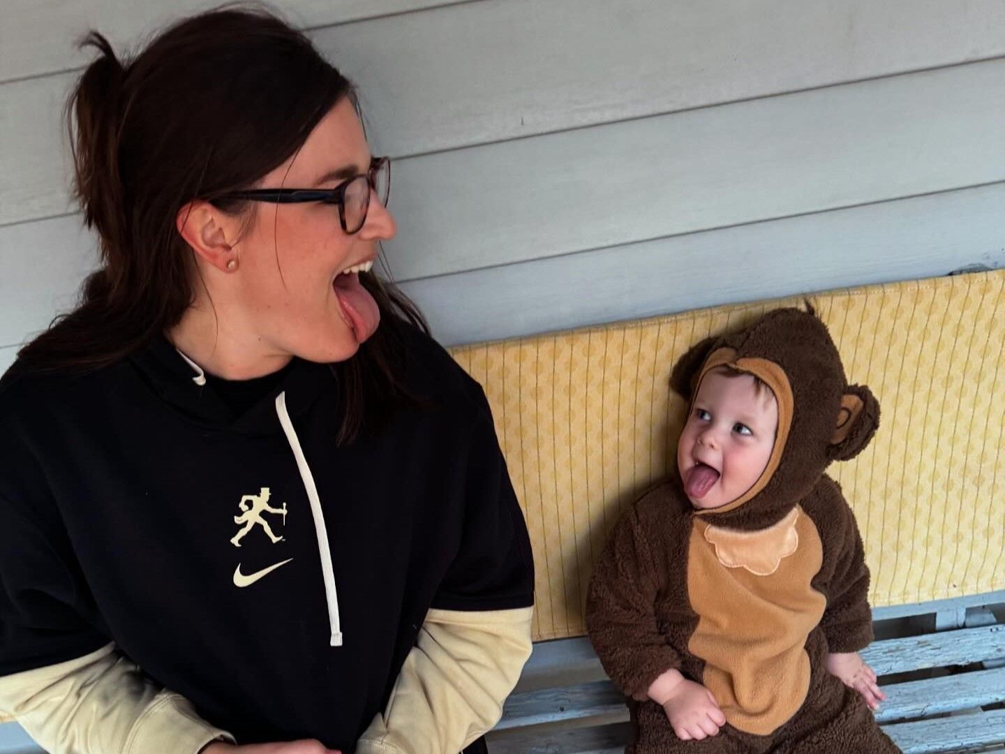 Person and baby in animal costume sitting on a porch bench, looking at each other with open-mouth smiles