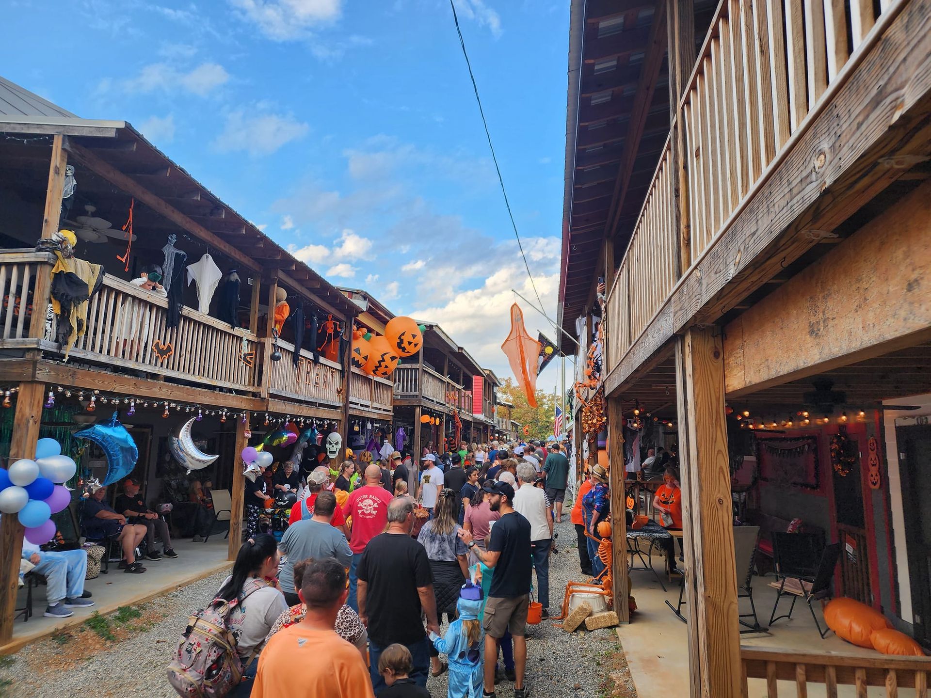 Crowded narrow street market between wooden buildings, with colorful flags and stalls under a blue sky