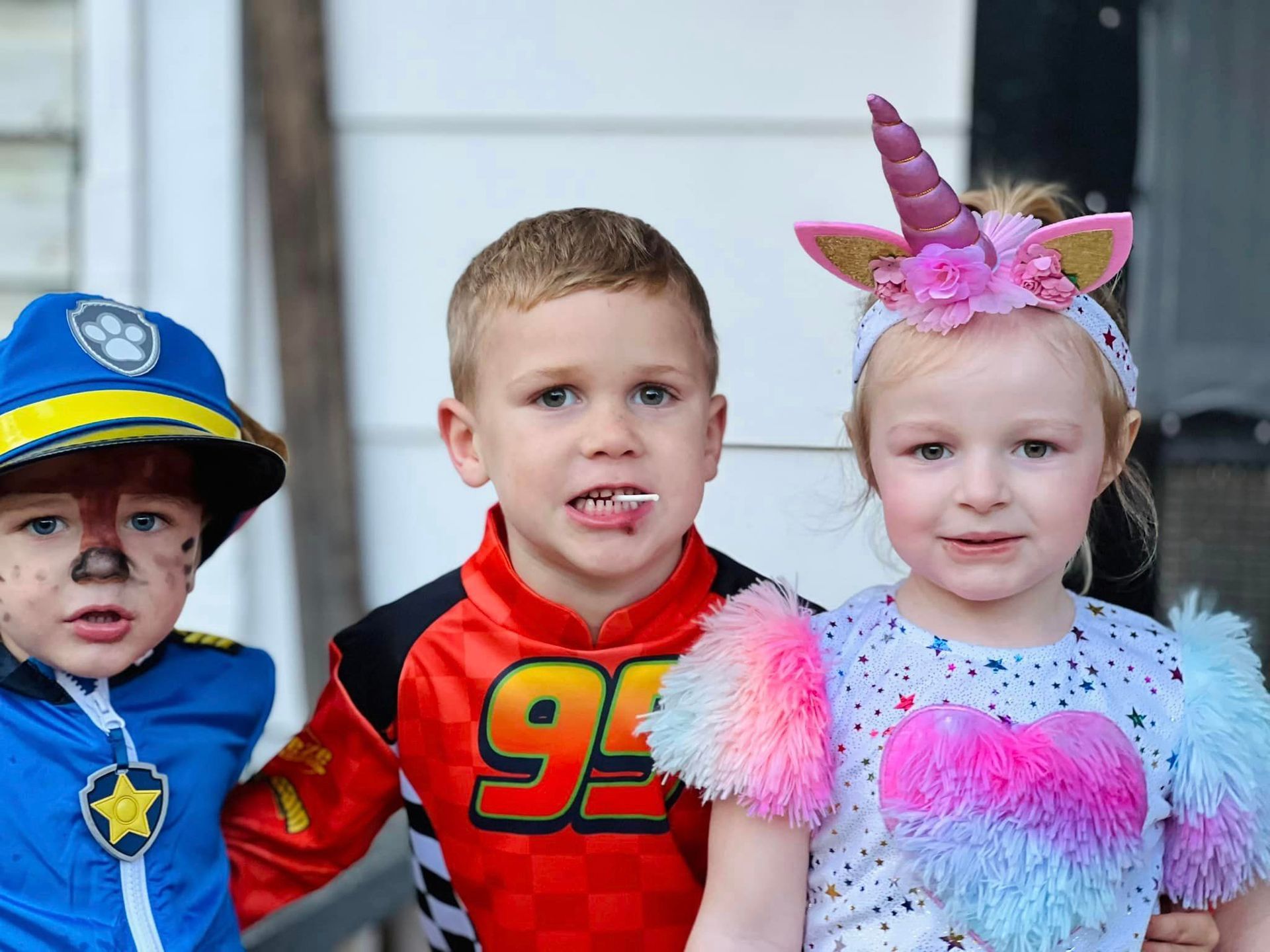 Three children in colorful costumes posing indoors, including a police outfit, a race car shirt, and a fairy costume.