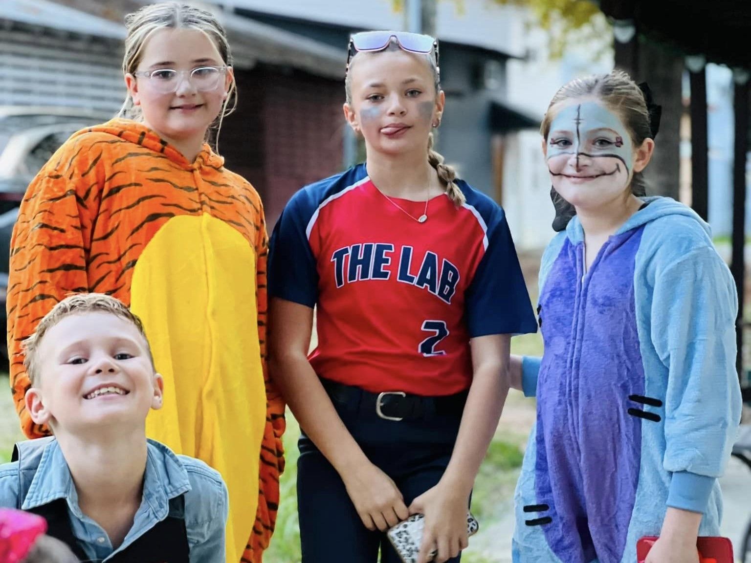 Four children in animal costumes posing outdoors, including a tiger, blue outfit, and red shirt.