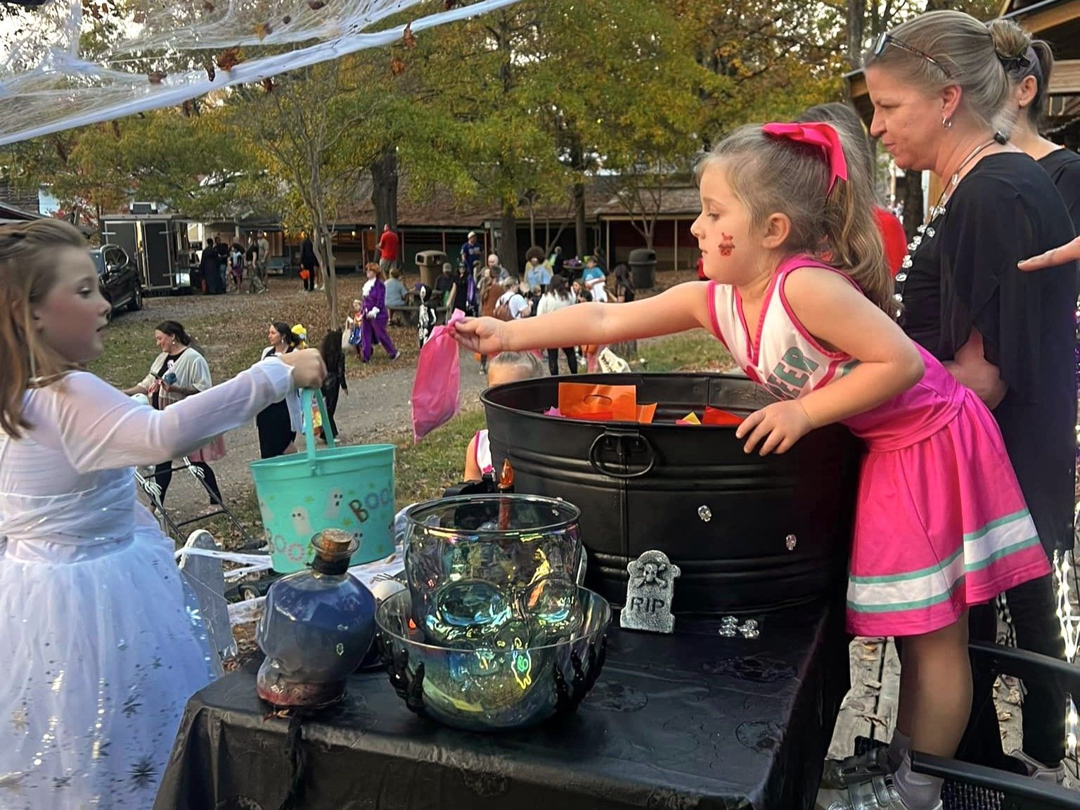 Children reaching into a black cauldron at an outdoor Halloween event with glass jars and decorations