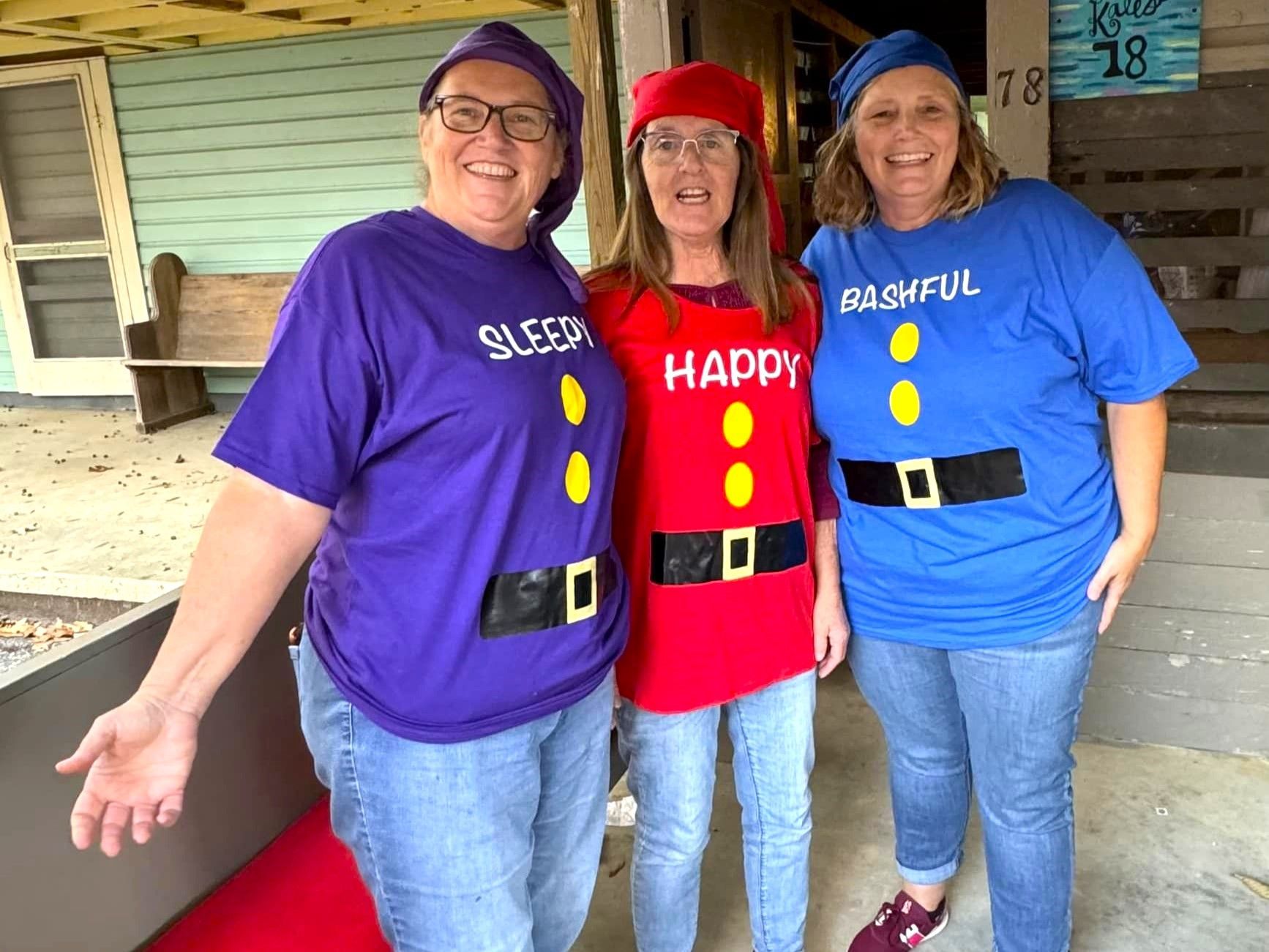 Three smiling people in colorful costumes standing on a porch, with painted shirts and matching hats.