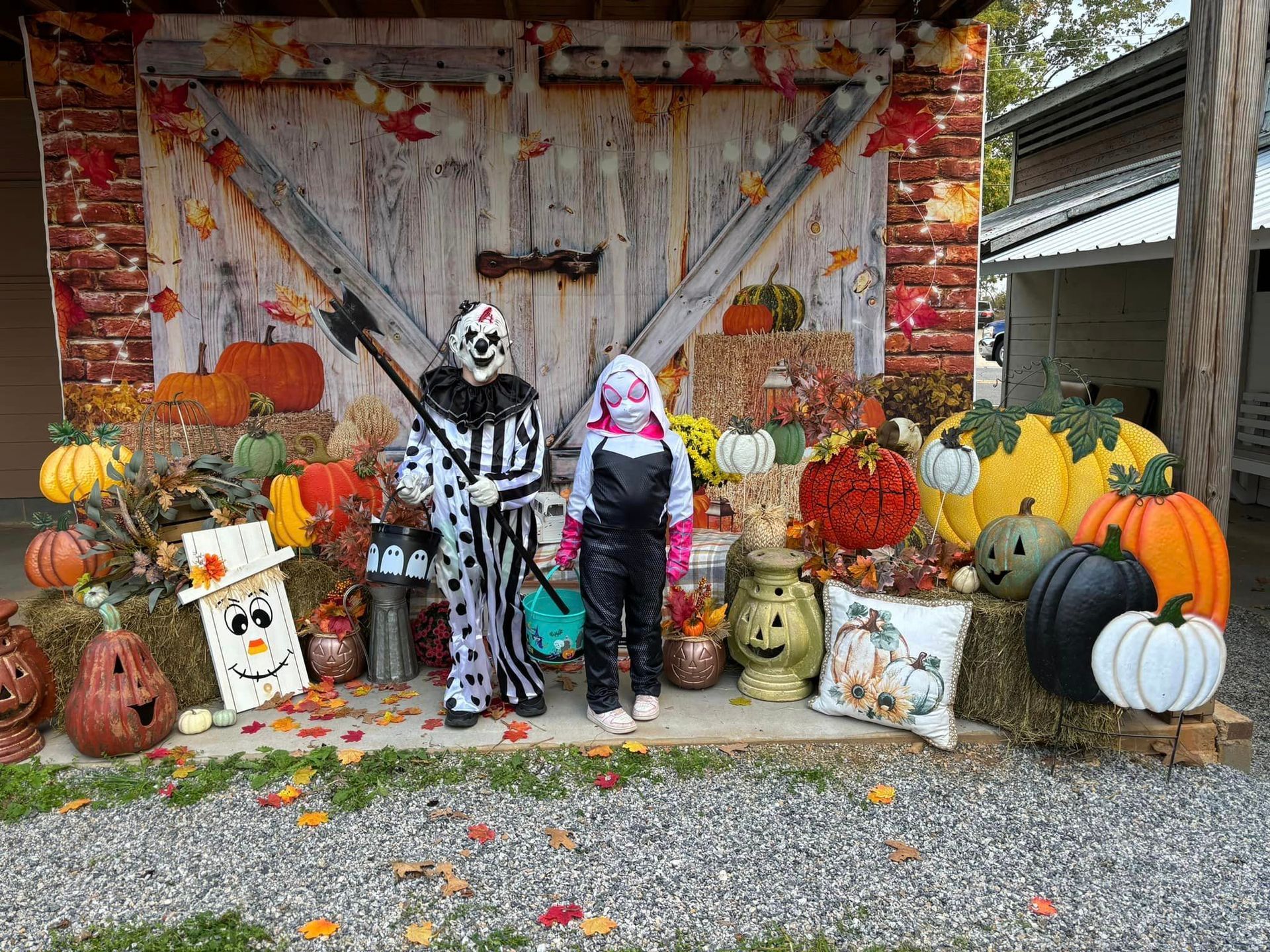 Halloween display with two skeleton figures amid pumpkins, hay bales, and fall decorations in front of a barn-like wall