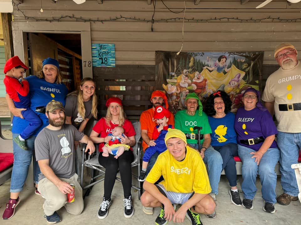 Group of people posing on a porch, wearing colorful hats and shirts, with one person in a yellow shirt kneeling in front
