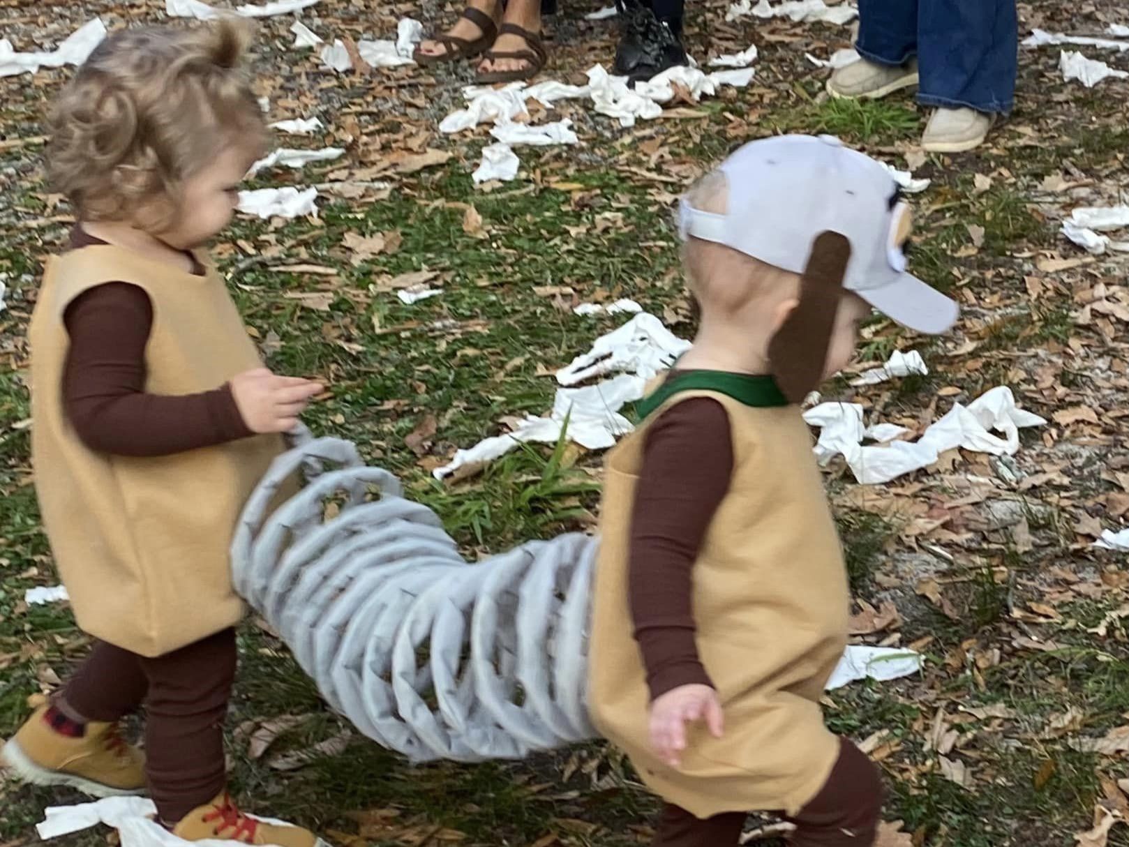 Two children in brown costumes with a gray tail-like tube, standing on grass scattered with white paper.