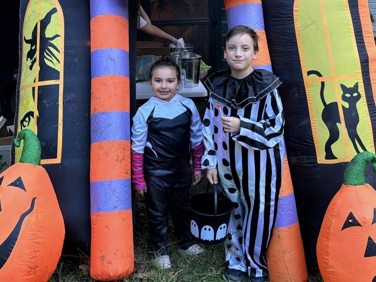 Two children in Halloween costumes standing by a spooky decorated entrance with pumpkins and a skull bucket.