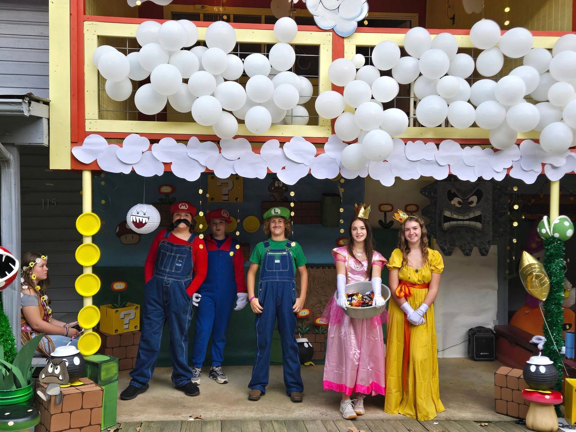 People standing under white balloon decorations at a storefront, some dressed in colorful costumes and holding flowers.
