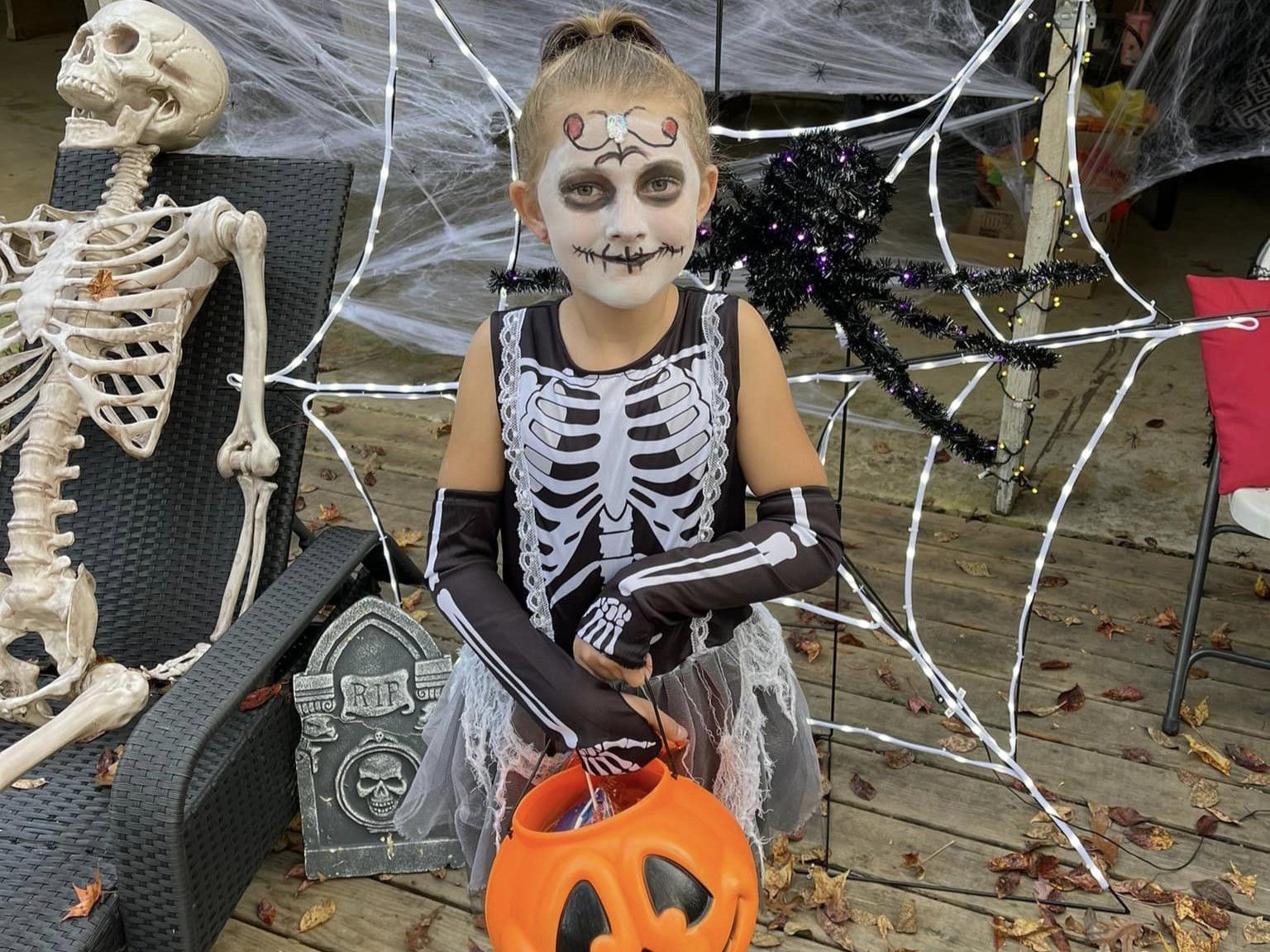 Child in skeleton costume with face paint, holding a pumpkin bucket beside Halloween skeleton decorations.
