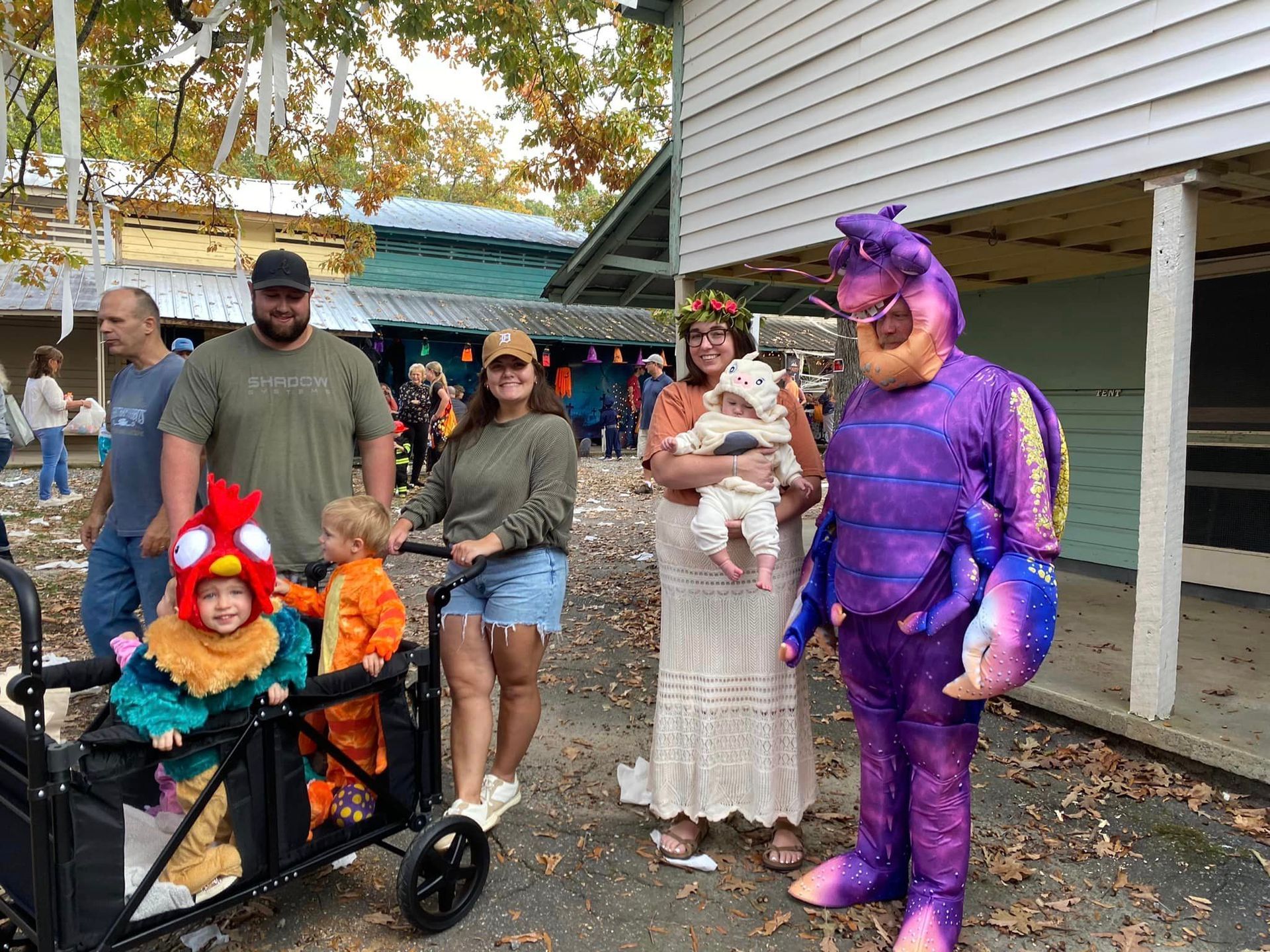 Group in Halloween costumes posing outdoors, including a purple dinosaur suit and a child in a stroller.