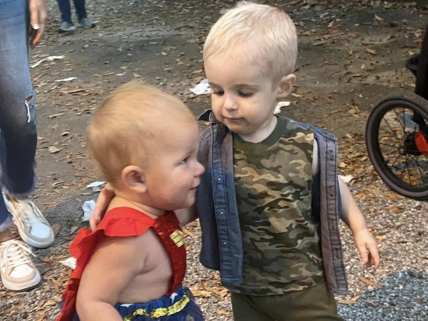 Two toddlers standing close together outdoors, one in a red outfit and one in a camouflage shirt.