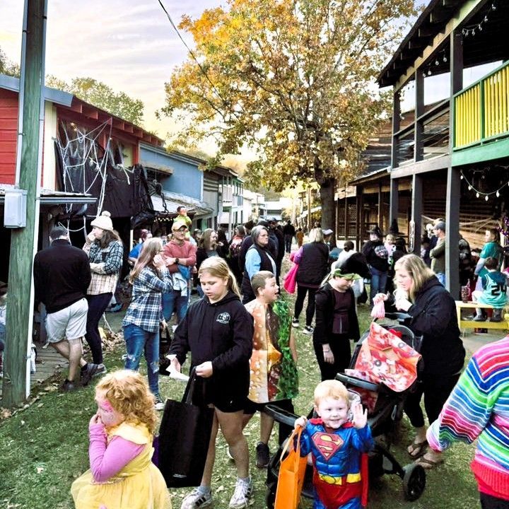 Crowded outdoor market or festival with people browsing beside wooden stalls under a large tree