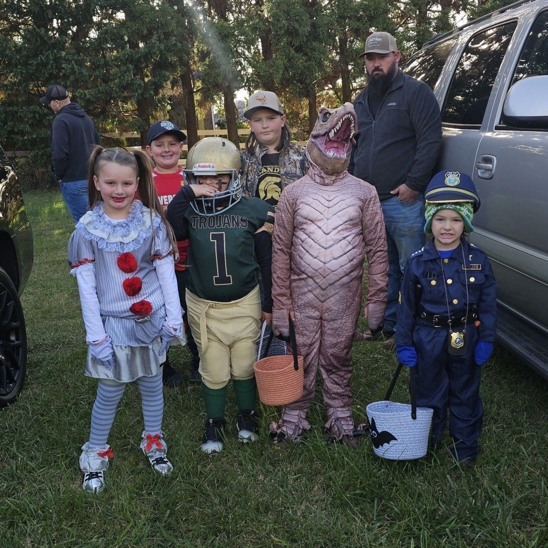 Children in costumes posing outdoors beside an SUV, with trees in the background.