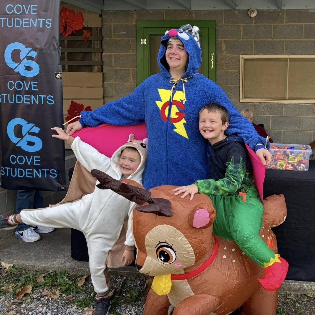 Three smiling people in colorful animal costumes sitting on a giant inflatable reindeer outside Cove Students