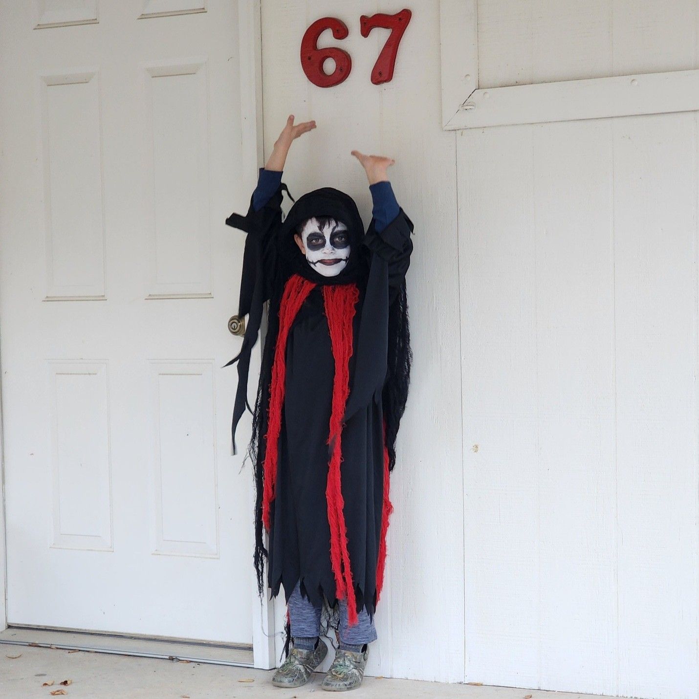 Child in black-and-red costume with face paint, standing with arms raised in front of a door marked 67.