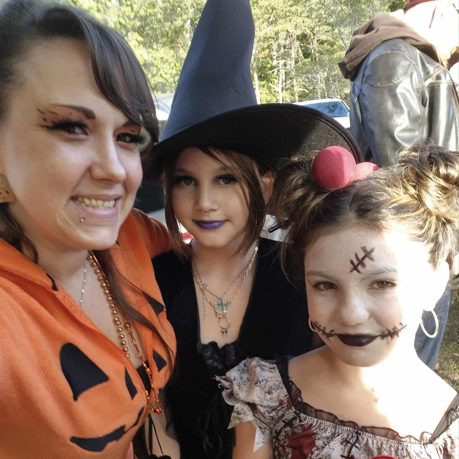 Three people in Halloween costumes pose outdoors, including a witch, pumpkin outfit, and stitched-face makeup.