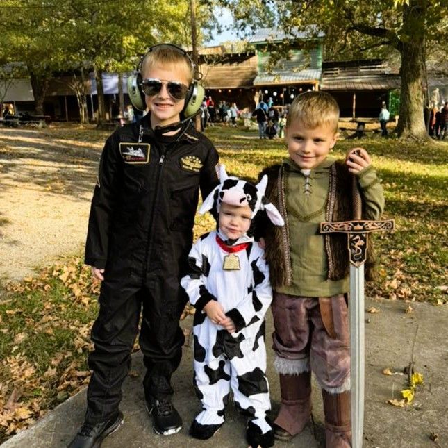 Three children in costumes standing outdoors on a sunny path, one dressed as a cow.
