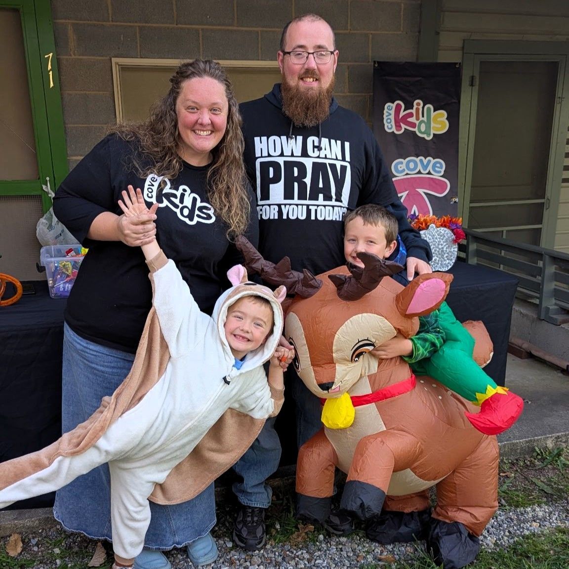Family in costume posing outdoors by a porch, including a dinosaur, cow, and horse, with a “COVID” sign.