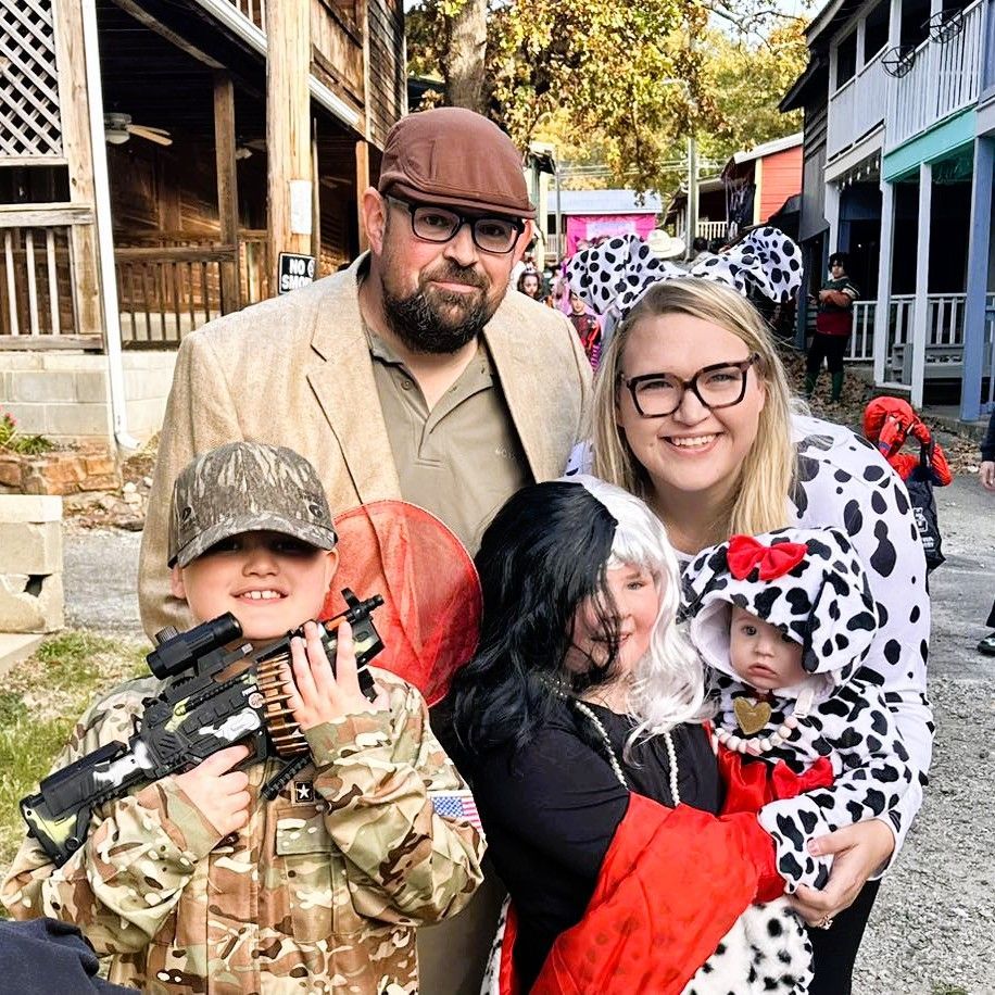 Family in costumes posing on a street, with a child holding a toy rifle and a red-cloaked child in front