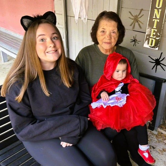 Three people on a porch, including a baby in a red hooded costume and a woman wearing black cat ears.