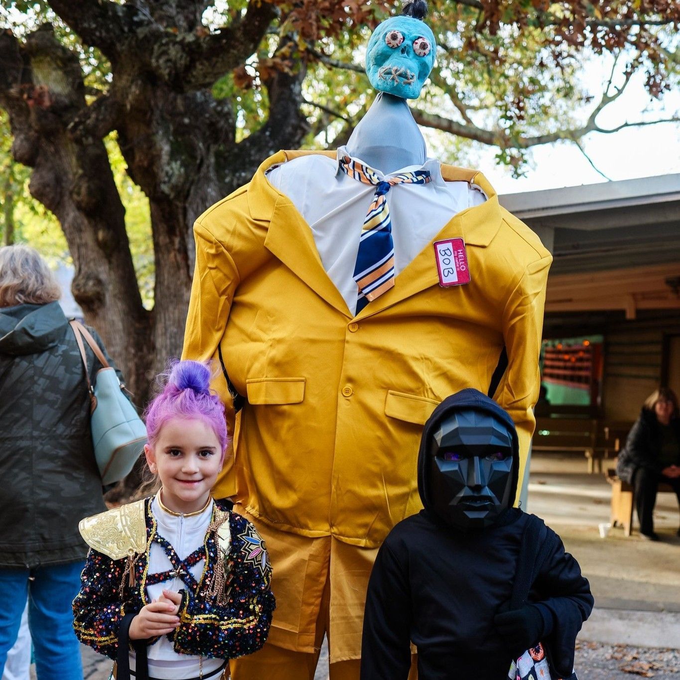 Two children in costumes posing with a giant blue-headed figure in a yellow suit outdoors