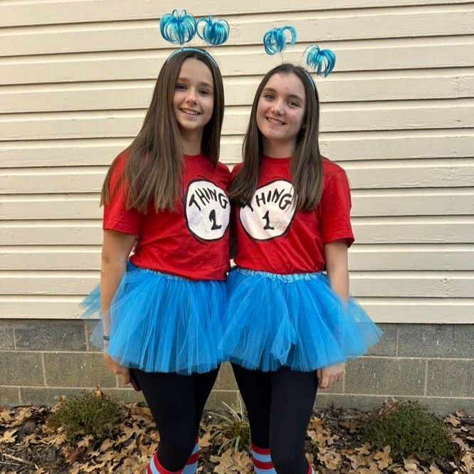 Two people in red “Thing 1” shirts and blue tutus posing in front of beige siding.