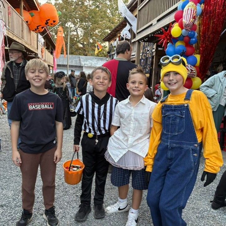 Four children in Halloween costumes standing on a decorated street with balloons and pumpkins.