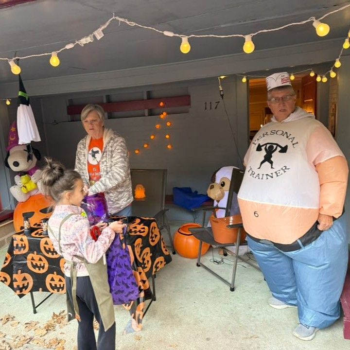 Halloween porch scene with jack-o'-lantern decor and two adults in costumes near a child in a purple cape