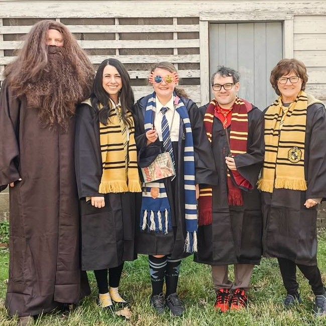 Five people in Hogwarts-style robes and scarves standing together outdoors, smiling in front of a wooden wall.