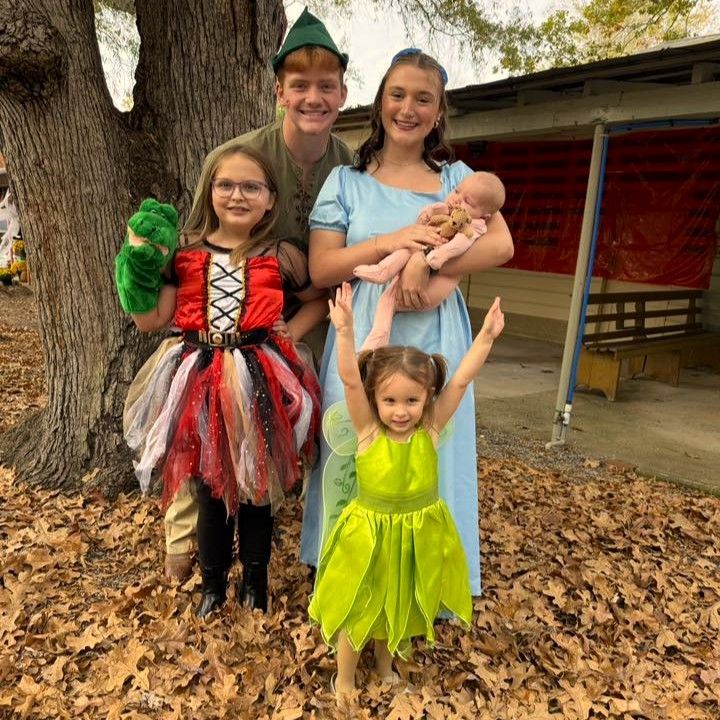 Costumed family posing outdoors under a tree, with two children and a baby in arms, one child in green dress raising both arms