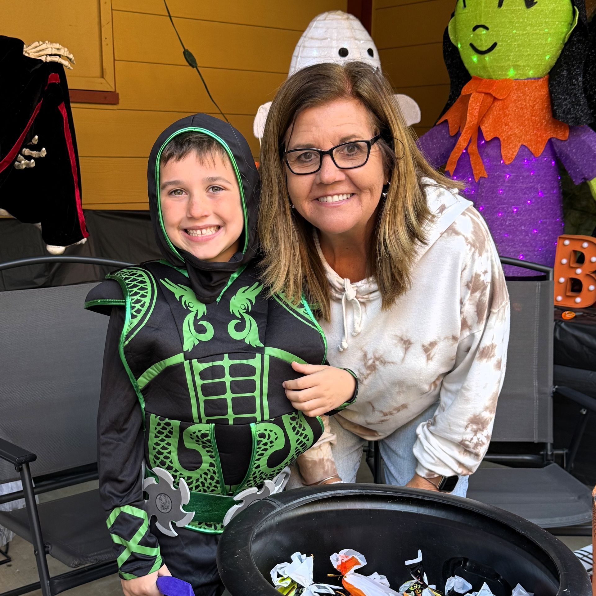 Smiling child in a black-and-green skeleton costume with an adult beside a Halloween display and candy bowl