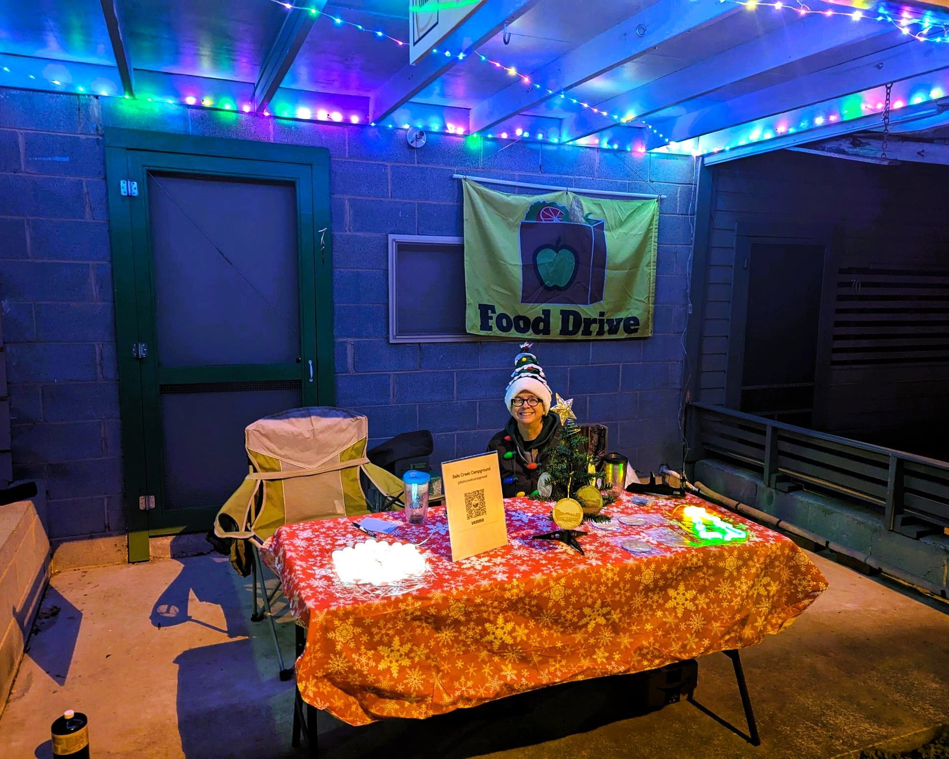 Food drive table under blue lights with a volunteer, donation boxes, and a banner in a dark outdoor setting